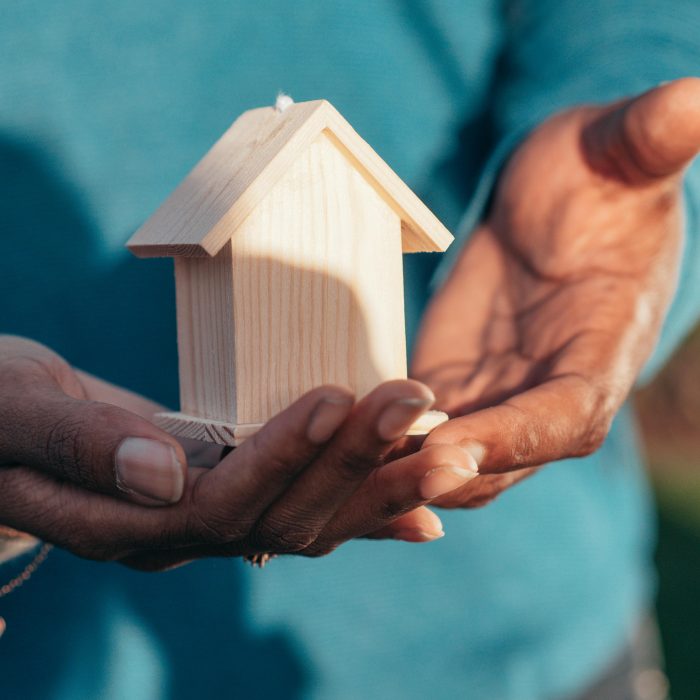 Close-up of hands holding a small wooden house model outdoors, symbolizing real estate investment.